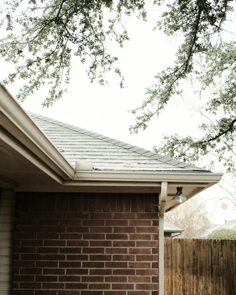 Corner of a red brick house with a grey shingle roof and white gutter under oak branches